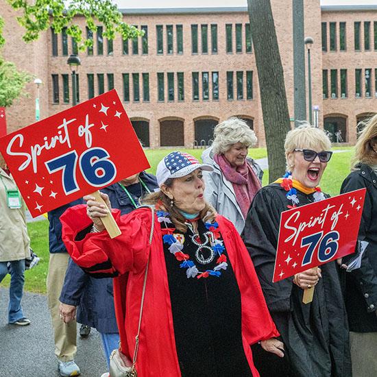 Two people in festive academic regalia walk outdoors on a campus pathway, each holding a bright red sign that reads “Spirit of ’76.”